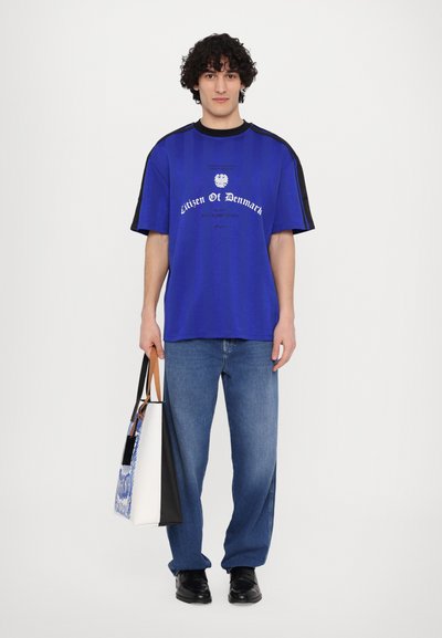 T-shirt bleu à manches courtes avec des accents noirs, comportant le texte "Citoyen du Danemark". Le modèle tient un sac fourre-tout multicolore et porte un jean bleu.