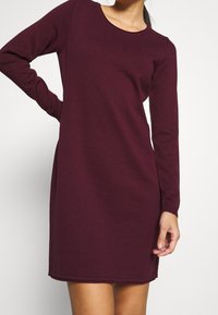 Burgundy stickad klänning med långa ärmar och rund halsringning. Har ribbat mönster, rak nederkant och en avslappnad passform.