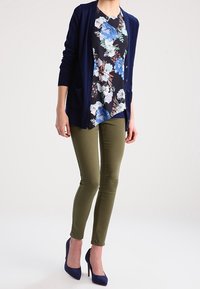 Blusa floreale blu sotto un cardigan navy, abbinata a pantaloni skinny verdi oliva e tacchi alti navy. Il cardigan ha tasche e bottoni.
