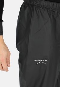 Zwarte waterdichte broek van lichtgewicht materiaal, met een elastische tailleband en een wit "SWEDEMOUNT" logo met bergafbeelding.
