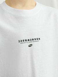 Balta medvilninė marškinėliai su apvaliu iškirpimu ir juodu spaudiniu, kuriame parašyta "JACK&JONES BROUGHT TO YOU BY ORIGINALS™" su metų "1990".