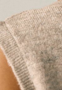 Beżowy sweter z dzianiny o fakturowanej powierzchni i ryfrowanych brzegach, ukazujący część ramienia i ręki. Widoczne rzadkie jasne odcienie.