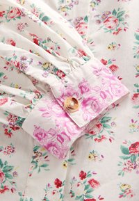 Material alb cu imprimeu floral multicolor, manșetă adunată cu model floral roz și închidere cu nasture maro marmorat.