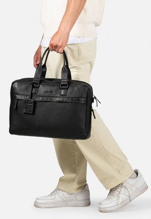 Malette en cuir noir avec deux poignées, surface texturée et branding embossé. Dispose de fermetures zippées et d'une étiquette amovible, portée par une personne.