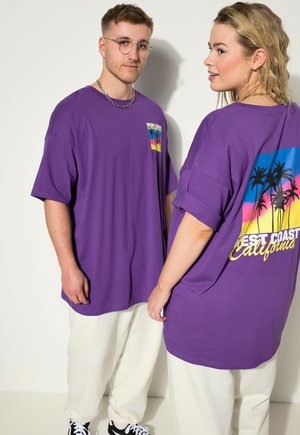 Due persone indossano magliette viola oversize con la scritta "West Coast California" e disegni di palme, abbinate a pantaloni bianchi e sneakers.