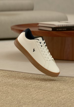 Witte sneaker met bruine zool en zwarte binnenvoering, zwevend boven een beige gestructureerd vloerkleed in een moderne woonkamer.