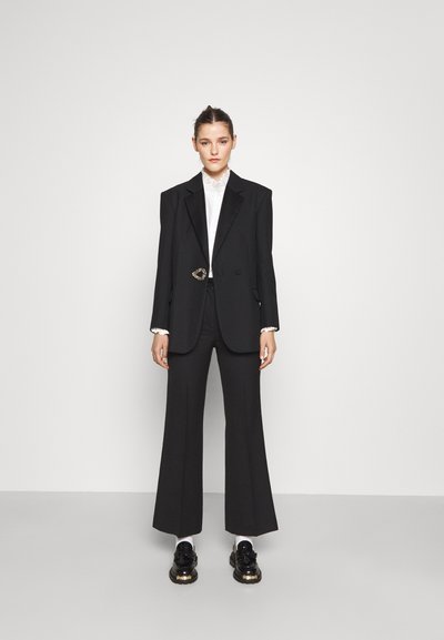 Sorte skræddersyet blazer med oversized skuldre og en rund dekorativ knap, parret med brede sorte bukser og blanke sorte sko.