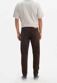 Pantaloni di cotone marrone con un design a gamba dritta, dotati di una toppa in pelle in vita, due tasche posteriori e scarpe blu. T-shirt grigia visibile.