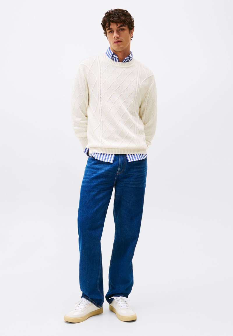 White knitted sweater with diamond pattern, layered over blue and white striped shirt, paired with blue jeans and white sneakers.