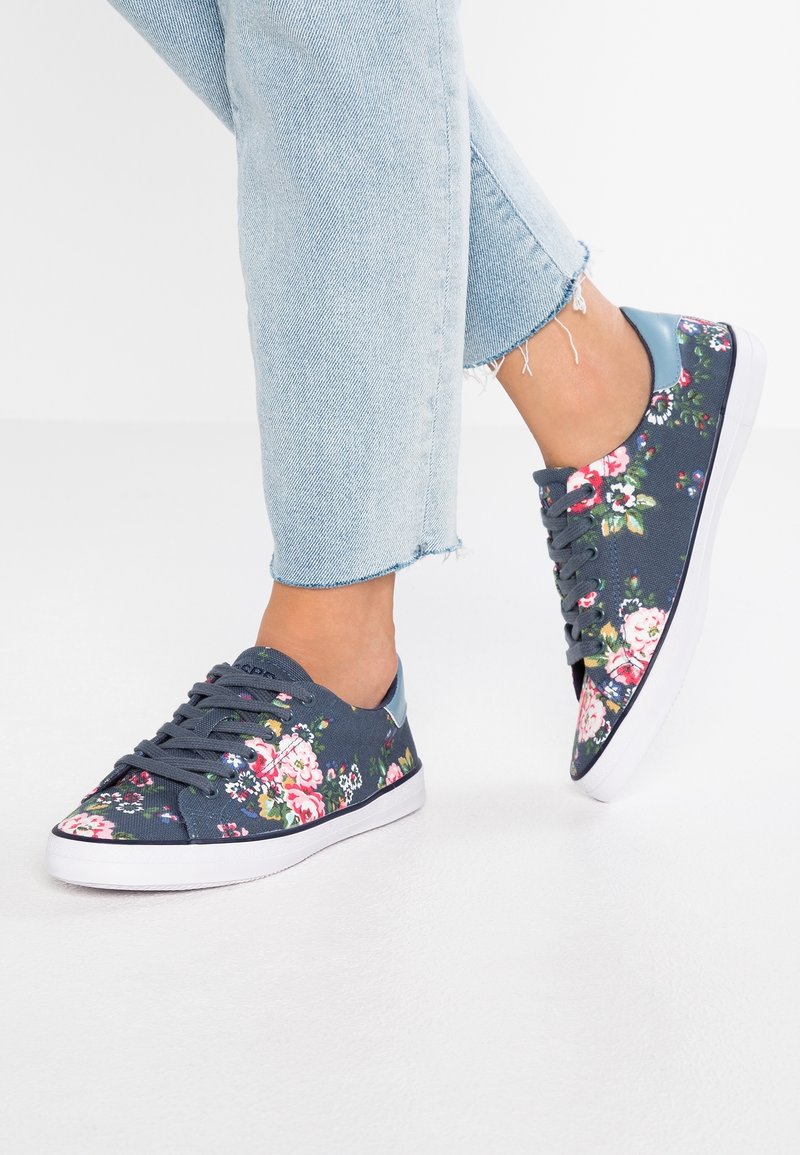 Personne portant un jean court bleu clair et des baskets bleu marine avec des motifs floraux roses et blancs, marchant sur une surface blanche.