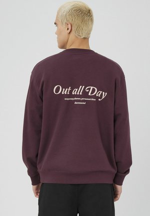 Homme aux cheveux blonds courts portant un sweat à capuche violet foncé avec "Sorti toute la journée" et un texte plus petit imprimé au dos, de dos.