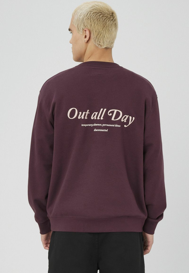 Homme aux cheveux blonds courts portant un sweat à capuche violet foncé avec "Sorti toute la journée" et un texte plus petit imprimé au dos, de dos.