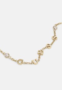 Aukso tono grandinėlė su pakabuku, ant kurio užrašytos raidės "G", "U", "E", "S", "S", papuoštos skaidriais apvaliais kristalais.