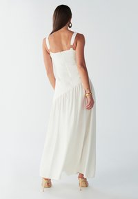 Bílé maxi šaty s tenkými ramínky a přiléhavým živůtkem, s uzavřením na zip a nařasenou sukní. Nošeny s páskovými podpatky.