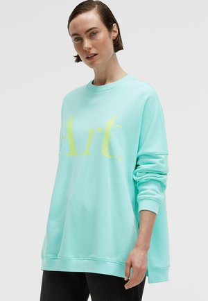 Woman with short brown hair wearing a loose light blue sweatshirt with yellow text "Art." and black pants, against a plain background.
