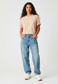 T-shirt beige clair avec des manches roulées, associé à un jean bleu clair à coupe ample et des baskets blanches. Tissu lisse et silhouette décontractée.