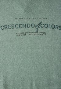 Tissu vert avec un texte en relief : "CRESCENDO COLORS" et des phrases en vert foncé, avec une surface texturée et un design décontracté.