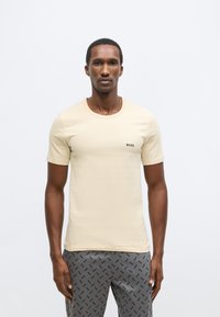 Maglietta beige a maniche corte con scollo tondo e logo nero "BOSS" sul petto, abbinata a pantaloni grigi con motivo e nome del marchio.