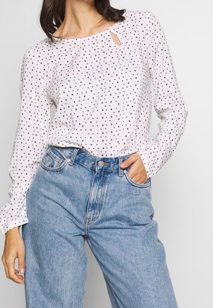 Blusa bianca a maniche lunghe con motivo a pois neri e dorati, scollatura rotonda con dettaglio a chiave, abbinata a jeans azzurri a vita alta.