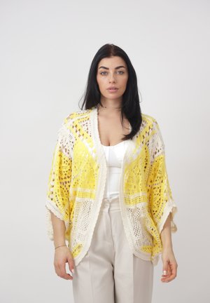 Woman with long dark hair wearing a white top, beige pants, and a yellow-white crochet cardigan standing against a plain light background.