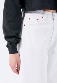 Jeans de mezclilla blanca con remaches de tono cobre y un ajuste de talle alto, combinados con un sudadera corta oscura de manga larga.