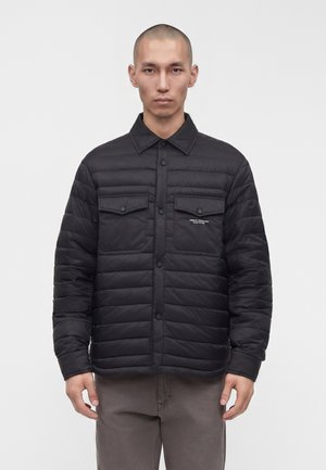Homme portant une veste matelassée noire avec des poches poitrine boutonnées et un pantalon gris foncé, debout devant un fond blanc uni.