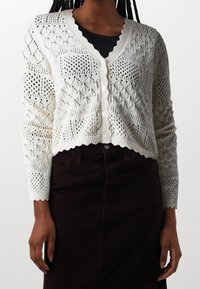 Biały sweter na guziki z dzianiny z wzorem diamentowym, falowanym dołem i mankietami, noszony na czarnym topie z zaokrąglonym dekoltem.