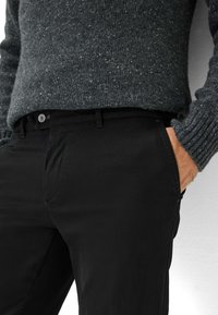 Pantaloni neri in un tessuto liscio, con chiusura a bottone e tasche laterali, abbinati a un maglione grigio scuro strutturato.