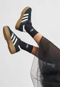 Black suede sneakers with white stripes, brown gum sole, and gold accents. Worn with black crew socks featuring a logo and a sheer tulle overlay.