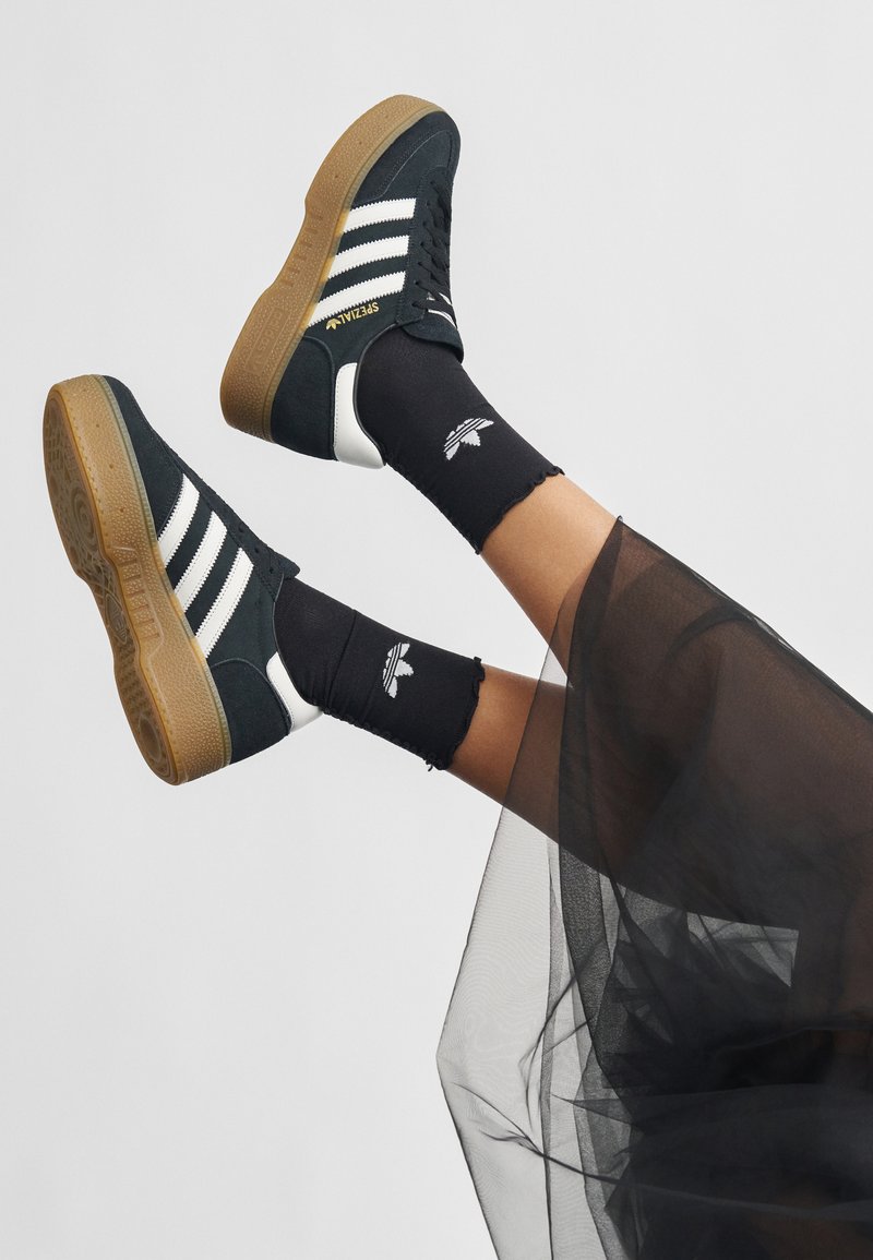 Black suede sneakers with white stripes, brown gum sole, and gold accents. Worn with black crew socks featuring a logo and a sheer tulle overlay.