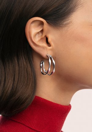 Primer plano de la oreja de una mujer que lleva pendientes de aro plateados retorcidos, con cabello oscuro y un cuello alto rojo visible.