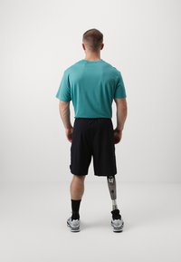 Atleta con una camiseta de manga corta color verde azulado y pantalones cortos negros, que presenta una pierna protésica y calcetines negros con zapatillas blancas. Fondo minimalista.