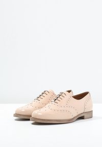 Chaussures en cuir verni beige avec des détails en brogue, bout arrondi, lacets et un talon en bois bas. Texture lisse sur l'ensemble.