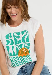 Camiseta sin mangas blanca con el texto ondulado verde "OLA DEL MAR", un sol naranja con dos aves y patrones de olas azules en la parte inferior.