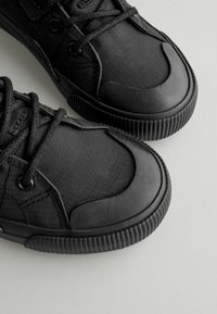 Schwarze Sneaker mit einem strukturierten Obermaterial aus Stoff mit Karomuster, gummierter Zehenkappe und genoppter Sohle. Die Schnürsenkel sind schwarz und besitzen verstärkte Ösen.