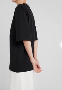 Černé tričko s krátkým rukávem, uvolněného střihu a kulatým výstřihem, s hladkou texturou a minimalistickým designem.