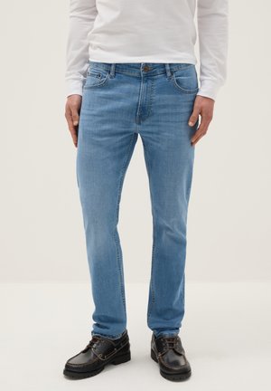 Hombre con vaqueros azul claro, camisa blanca de manga larga y zapatos marrón oscuro con cordones, de pie contra un fondo liso.