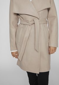 Beige kappa med bred krage, självbunden bälte och två sidofickor. Tillverkad av mjukt, strukturerat tyg. Vit detalj vid ärmslut.