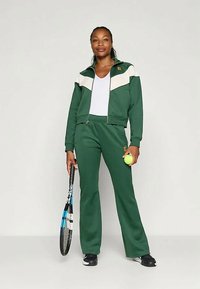 Grön träningsoverall med en vit rand, zip-up jacka och byxor med elastisk midja. Håller en tennisracket och boll. Slät textur, sportig design.