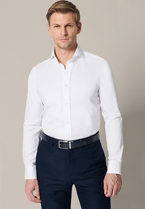 Mężczyzna ubrany w białą koszulę z długim rękawem, włożoną do ciemnogranatowych spodni z czarnym paskiem, stoi na jednolitym jasnym tle.