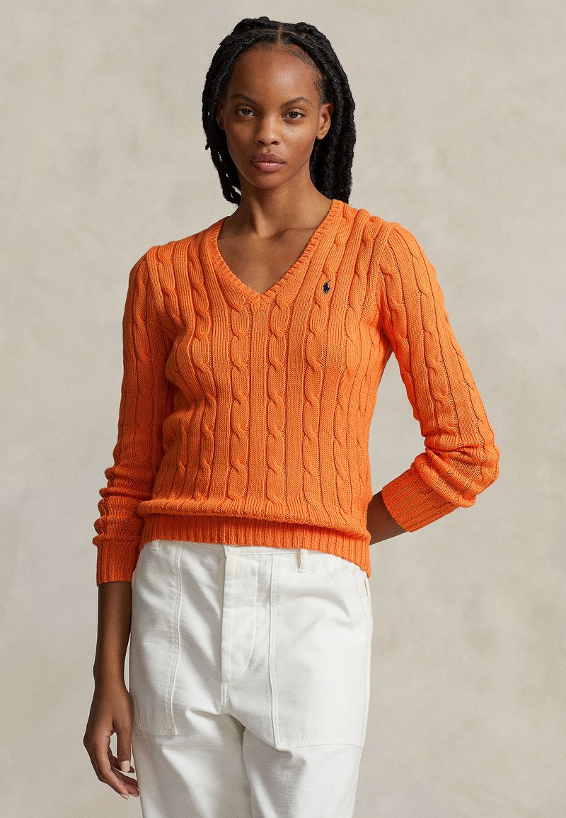 Pull en maille orange avec un col en V et des manches longues, présentant un design ajusté et des poignets côtelés. Pantalon blanc en arrière-plan.