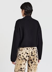 Schwarzer, kürzer geschnittener Pullover mit einem gerippten Kragen und langen Ärmeln, kombiniert mit beigen Hose mit gepunktetem Muster. Texturiertes Strickgewebe.