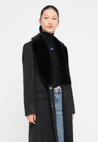 Mustra mustriga must coat koos musta kunstkarusnahast kraega, millel on kaks eesmist taskut ja hõbedased nööbid, kantud musta ribakraega turtleneck'i üle.