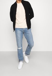 Černá bunda na zip přes světle béžové tričko, doplněná světle modrými džínami s opotřebovaným vzhledem a bílými teniskami. Hladké materiály, neformální střih.