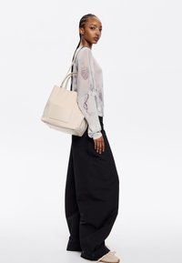 Bolso de cubo de cuero beige con asas dobles, que presenta un logo en relieve. Modelo luciendo una blusa transparente con estampado y pantalones de pierna ancha negros.