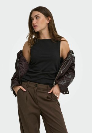 Jonge vrouw die een zwart mouwloos topje, een bruine broek en een gedeeltelijk gedragen donkerbruine leren jas draagt, kijkt naar links met haar handen in haar zakken.