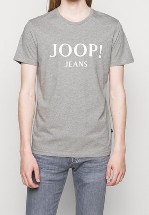Personne portant un T-shirt gris à manches courtes avec « JOOP! JEANS » imprimé en blanc et un jean en denim bleu clair.
