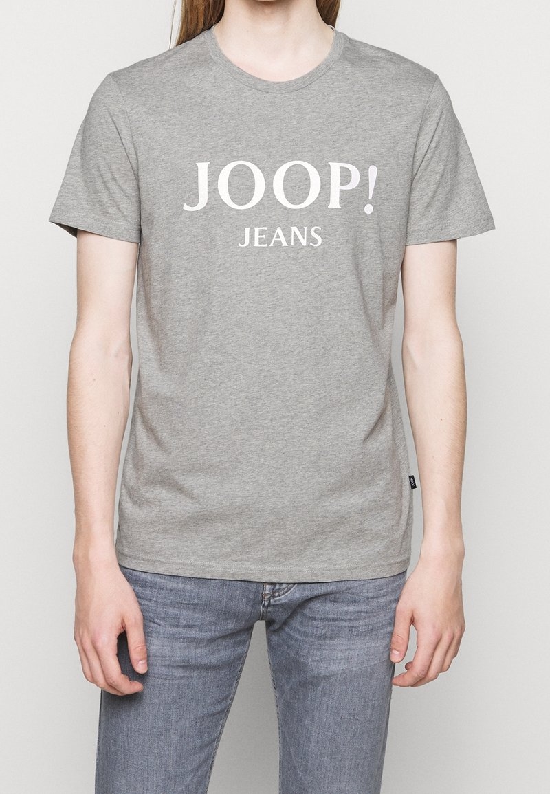 Person trägt ein graues kurzärmeliges T-Shirt mit dem Aufdruck "JOOP! JEANS" in Weiß und hellblauen Jeans.
