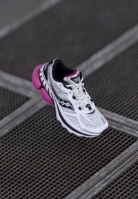 White athletic shoe with pink accents, mesh upper, and a thick sole. Features include a padded collar, lace-up design, and logo detailing.