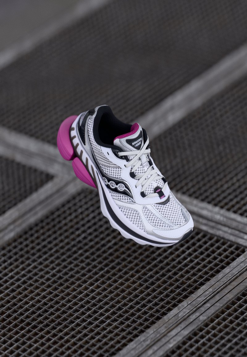 Zapatilla deportiva blanca con detalles en rosa, parte superior de malla y suela gruesa. Las características incluyen un collar acolchado, diseño con cordones y detalle de logo.